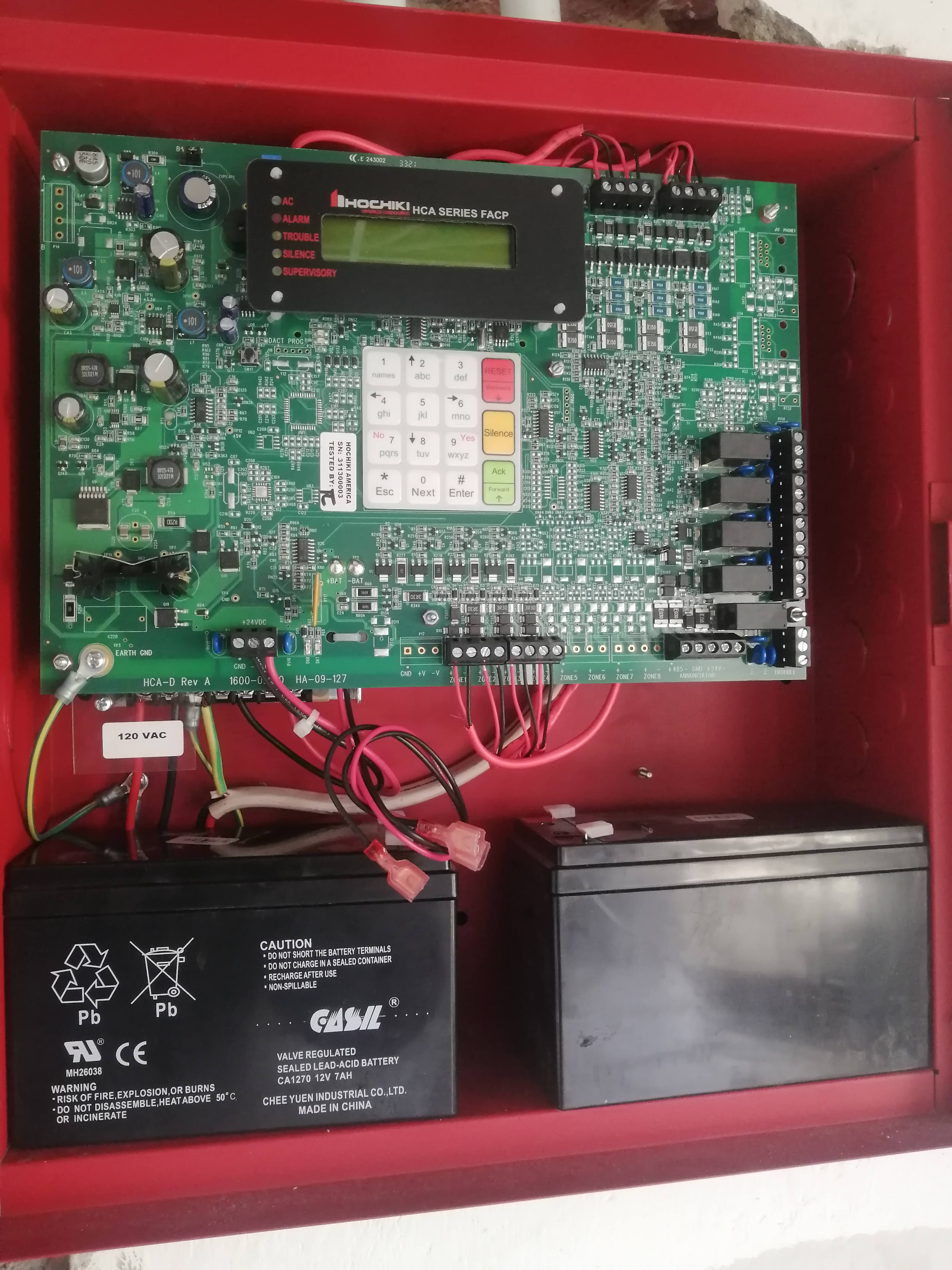 Red fire alarm control panel containing a green circuit board and two black batteries.